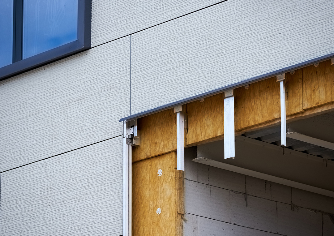 Close-up view of a building under construction, showing the exterior wall with layers of siding, insulation, and structural framework exposed. The image highlights the details of modern construction techniques and materials used for energy efficiency and durability.